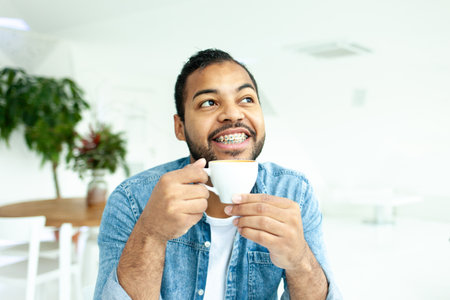cheerful african american man with braces holds a cup of coffee and plans and dreams in a white cafe, pensive man in a denim shirt drinks coffee in the morning and imaginesの写真素材