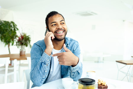 cheerful african american man with braces talking on the phone in a white cafe, a man in a denim shirt smiles and communicates on a mobile phone in a restaurantの写真素材