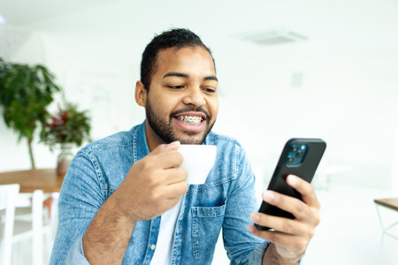 cheerful african american man with braces holds a cup of coffee and a smartphone in a white cafe, a man in a denim shirt drinks coffee in the morning and uses a mobile phone onlineの写真素材