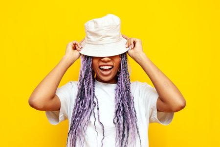 young african american woman with hipster colored dreadlocks wears hat on yellow isolated background, girl with unique purple hairstyle hides and covers her eyes with hatの写真素材