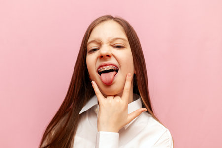 little child girl with braces showing rock gesture with hands and tongue on pink isolated background, crazy teenager showing heavy music symbolの写真素材
