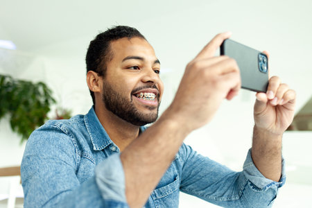 cheerful african american man with braces uses a smartphone and takes pictures in a white room, a man in a denim shirt holds a mobile phone horizontally and watches a videoの写真素材