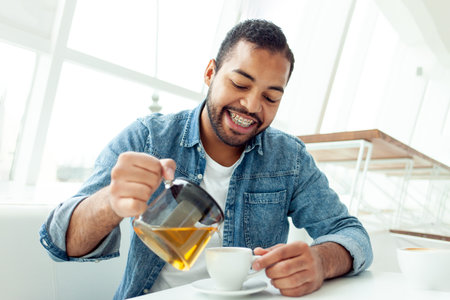 happy african american man with braces holds a teapot and pours tea into a cup in a white cafe, the man smiles and has breakfast in a restaurantの写真素材