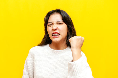 young dissatisfied asian girl with braces threatens and shows fist on yellow isolated background, angry korean girl swears and quarrelsの写真素材