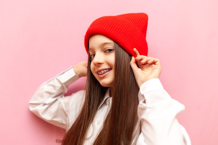 cheerful little teenage girl with braces puts on a warm red hat on a pink isolated background, child smiling in a winter knitted hatの写真素材