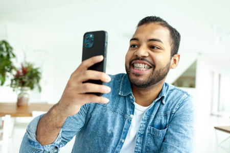 cheerful african american man with braces uses smartphone and takes pictures in white room, man in denim shirt holds mobile phone and photographingの写真素材