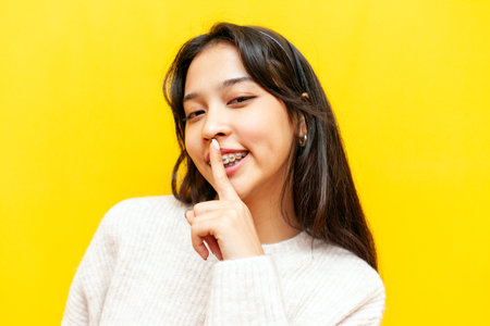 young cheerful asian woman with braces shows finger near lips and gesture of secret on yellow isolated background, korean girl shows silence and forbids speakingの写真素材