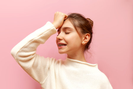 excited teenage girl is worried and shows a facepalm on a pink isolated background, an unhappy child makes a mistake and shows a fail gestureの写真素材