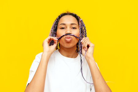 young african american woman with dreadlocks shows a mustache from her hair on a yellow isolated background, a girl with a unique hairstyle and colored braids makes faces and jokesの写真素材