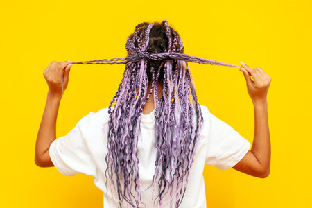 young african american woman with dreadlocks holding her hair in her hands on a yellow isolated background, girl with a unique hairstyle and colored braids smilingの写真素材