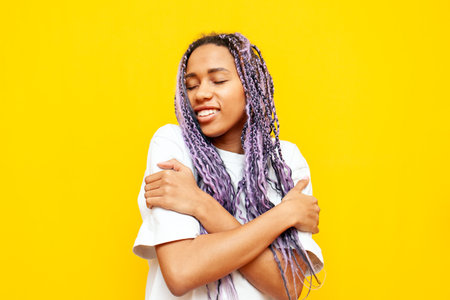 young african american woman with dreadlocks hugs herself over yellow isolated background, girl with unique hairstyle and colored braids loves herselfの写真素材
