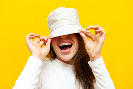 cheerful woman with braces smiling and screaming covering her face with a hat on a yellow isolated background, excited girl showing a dental smile with bracesの写真素材
