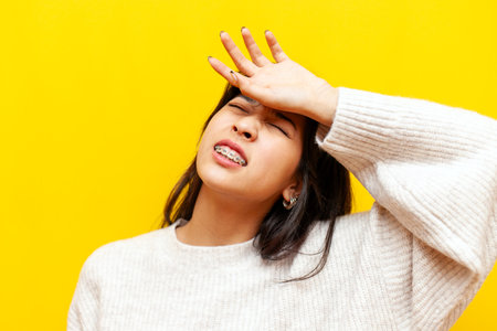 young tired asian woman with braces suffers and is sad on a yellow isolated background, korean unhappy girl wipes her forehead with her hand and shows dissatisfactionの写真素材