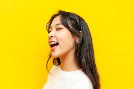 young asian woman with braces winks and smiles on yellow isolated background, korean girl hints and clicks her eyeの写真素材