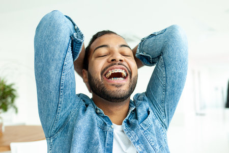happy african american man with braces laughing in a white cafe, relaxed man in a denim shirt holds...