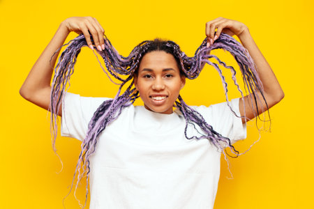 young african american woman with dreadlocks holding her hair in her hands on a yellow isolated background, girl with a unique hairstyle and colored braids smilingの写真素材
