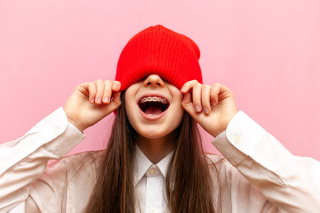 scared little teenage girl with braces hides behind a warm red hat and screams on a pink isolated background, the child puts on and closes his eyes with winter clothesの写真素材
