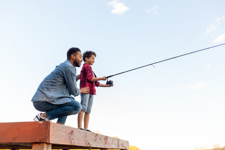African American man and child sitting on a wooden pier holding fishing rods on the river, dad teaching his son to fish in the lake, family resting and relaxing on the weekendの写真素材