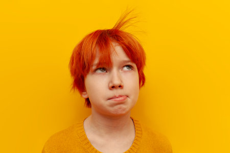 tired red-haired teenage boy is sad and blows on his hair on yellow isolated background, unhappy child with orange hairstyle is bored and disappointedの写真素材