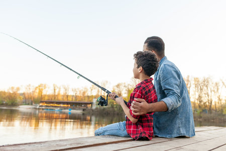 African American man and child sitting on a wooden pier holding fishing rods on the river, dad teaching his son to fish in the lake, family resting and relaxing on the weekendの写真素材