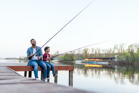 african american man and child holding fishing rods and fishing in the river, dad and son sitting on a wooden pier and fishing on the lake at sunsetの写真素材