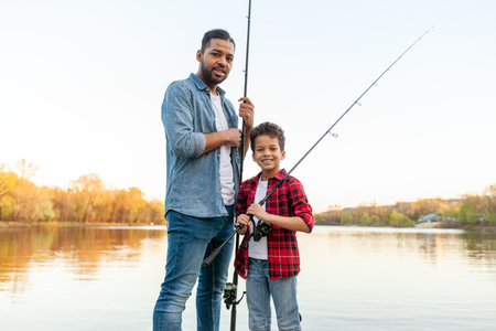 African American man and child sitting on a wooden pier holding fishing rods on the river, dad teaching his son to fish in the lake, family resting and relaxing on the weekendの写真素材