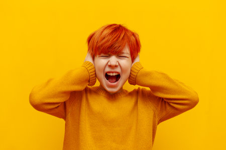 scared red-haired teenage boy screaming in stress and covering his ears on yellow isolated background, irritated angry child with orange hairstyle in panic and depressionの写真素材