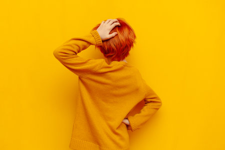 puzzled red-haired teenage boy thinks and chooses on a yellow isolated background, confused child with an orange hairstyle remembers and scratches the back of his head, rear viewの写真素材