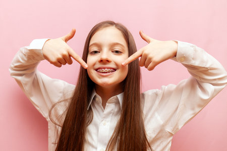 cheerful teenage girl with braces points with finger to the smile on pink isolated background, child...