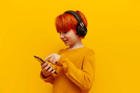 red-haired child boy in headphones listens to music and uses a smartphone on a yellow isolated background, a teenager with an orange hairstyle in black headphones chooses songs onlineの写真素材