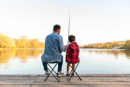 African American man and child sitting on a wooden pier holding fishing rods on the river, dad teaching his son to fish in the lake, family resting and relaxing on the weekendの写真素材