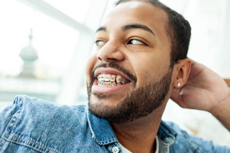 happy african american man with braces laughs and looks out the window in a white cafe, relaxed man in a denim shirt dreams and smilesの写真素材