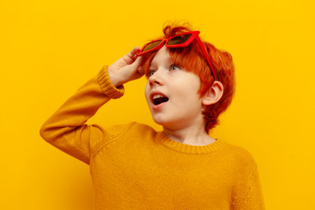 shocked red-haired child boy in sunglasses looking up at the sun with open mouth on yellow isolated...