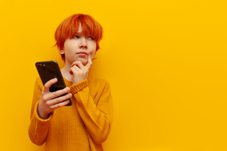 pensive red-haired child boy using smartphone and planning on yellow isolated background, puzzled...