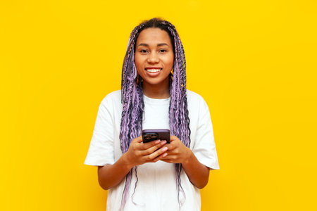 cheerful african american woman with colored dreadlocks using smartphone on yellow isolated background, hipster girl with purple braids and unique hairstyle holding mobile phoneの写真素材