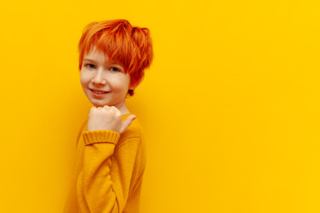 red-haired teenage boy points with his hands back on a yellow isolated background, a cheerful child with an orange hairstyle shows and advertises copy space behindの写真素材