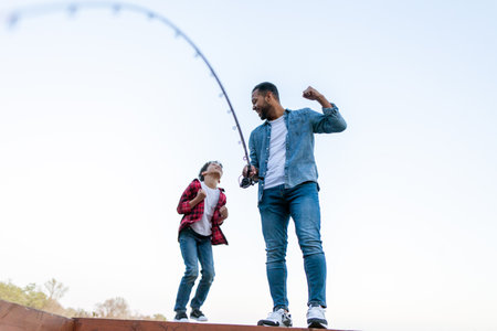 An African American father and his young son are celebrating their success on a wooden pier. The father, in a blue shirt and jeans, holds a fishing rod with a fish on the line, raising his fist in victory, while the son, in a red plaid shirt and jeans, jumps with excitement and joy. Both are delighted with their catch, with a clear sky and a lake in the backgroundの写真素材