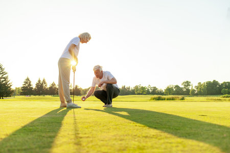elderly couple seniors playing golf on golf course at sunset, old man and woman practicing with golf clubs in nature, husband and wife in white uniform actively relaxingの写真素材