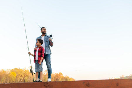 An African American father and his son stand on a wooden pier by a lake with fishing rods. The...