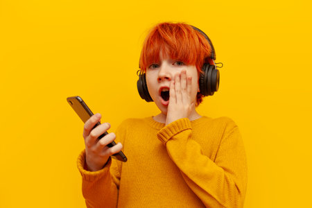 shocked red-haired child boy in headphones listens to music and uses smartphone on yellow isolated background, surprised teenager with orange hairstyle chooses online in amazementの写真素材