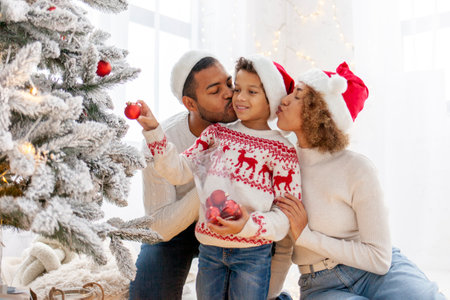 African American family in Santa hat celebrate New Year together at home and decorate Christmas tree, teenage boy celebrates New Year with mom and dad and holds red toyの写真素材