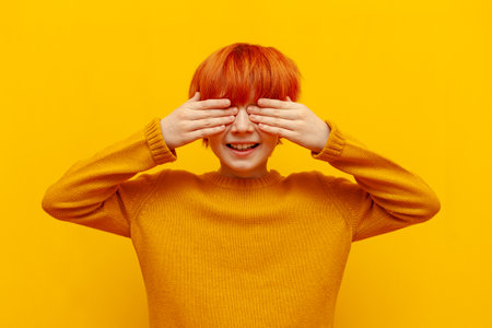 cheerful red-haired child boy closes his eyes with his hands and smiles on yellow isolated background, excited teenager with orange hairstyle does not look and waits for gift and surpriseの写真素材