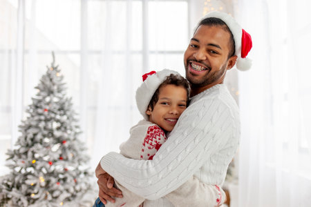 african american teenage boy in santa hat hugs dad at home for christmas, man wishes son happy new year and celebrates togetherの写真素材