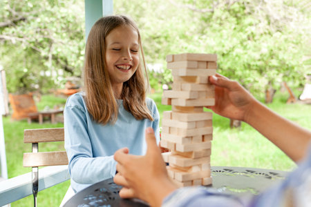 girl in a blue sweater watches with a smile as her friend carefully pulls a block from the middle of a tower of wooden cubes. Outdoor games bring joy and fun to children.の写真素材