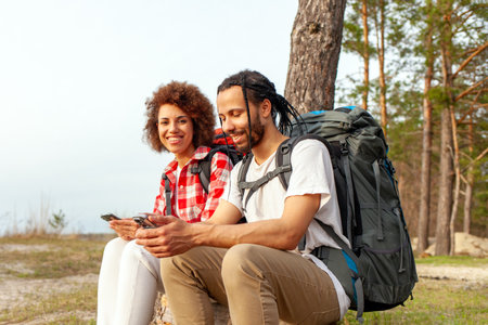 young african american couple hiking with backpacks sitting and using smartphones online in forest...