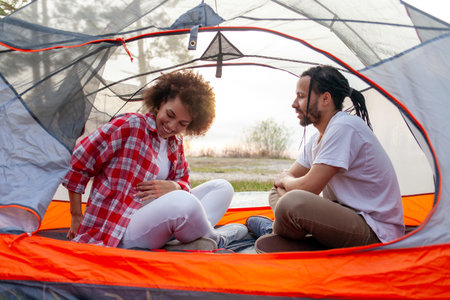 young african american couple tourists with camping equipment and backpacks sitting in a tent in the woods and talking, man and woman camping relaxing and talkingの写真素材