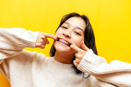 young crazy asian girl with braces stretches smile with fingers on yellow isolated background, korean girl shows wide smile and teethの写真素材