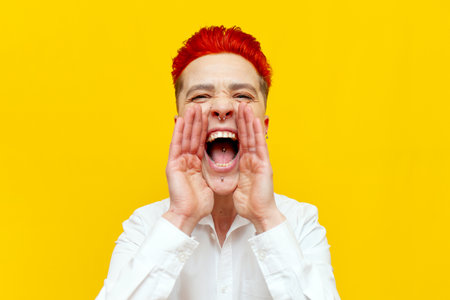 young short-haired red-haired girl announces loudly and speaks on a yellow isolated background, hipster woman with piercing screams with her mouth openの写真素材