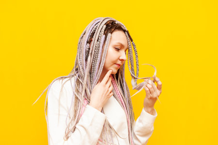 young girl with colored dreadlocks holds damaged hair and thinks on yellow isolated background,...