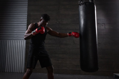 young strong african american boxer in red gloves hits punching bag, muscular athlete trains punch in dark gym and does sports on black backgroundの写真素材